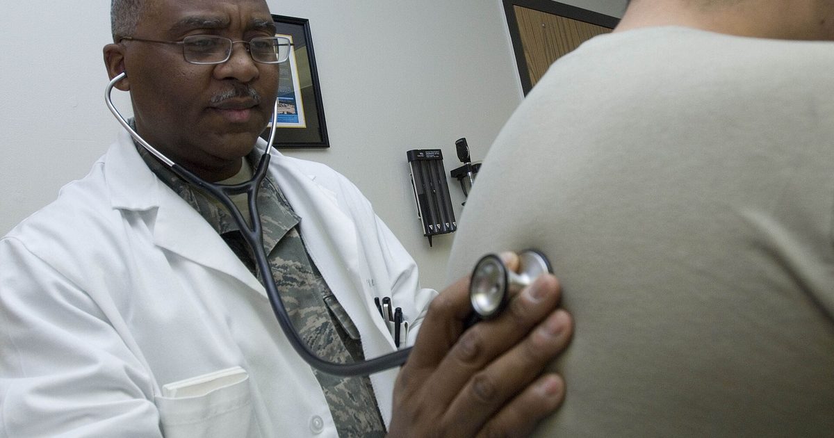 A physician in a white coat holds a stethoscope to an adult patient's bare back during an exam