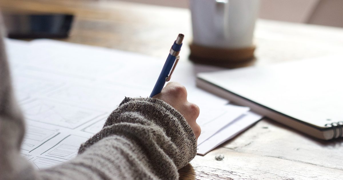 Hand holding a pen over printed pages on a wooden desk, with a mug and laptop nearby