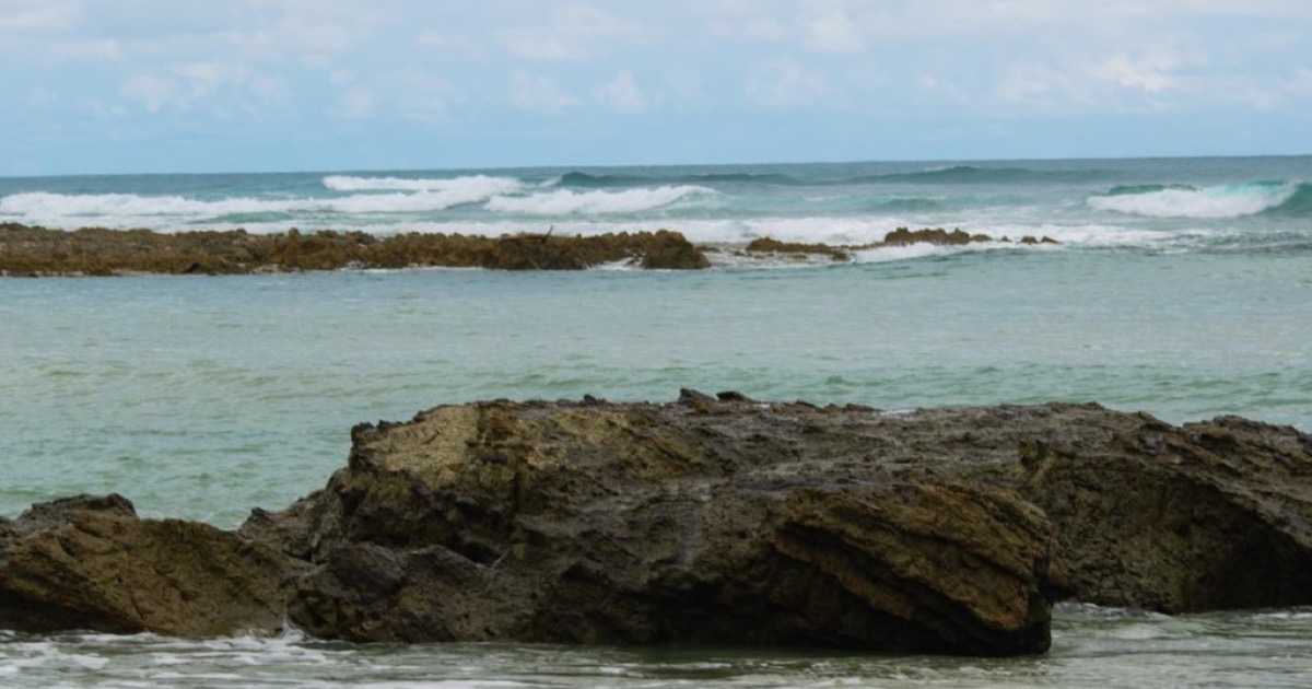 Waves breaking over a low rocky reef in shallow turquoise water under a pale, cloudy sky