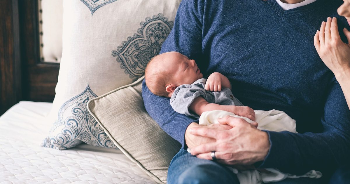 An adult in a blue sweater cradles a swaddled newborn while sitting on a bed with patterned pillows