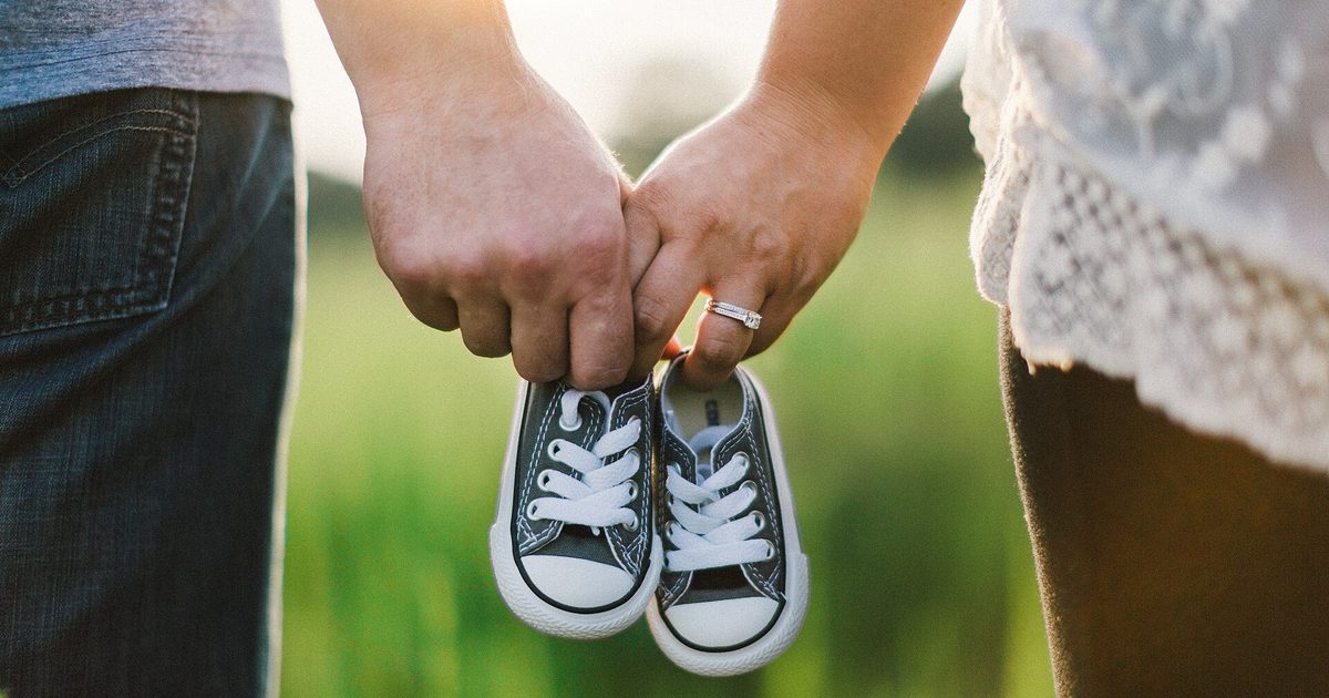 Two parents hold hands in a sunlit field, a pair of tiny baby sneakers dangling between them