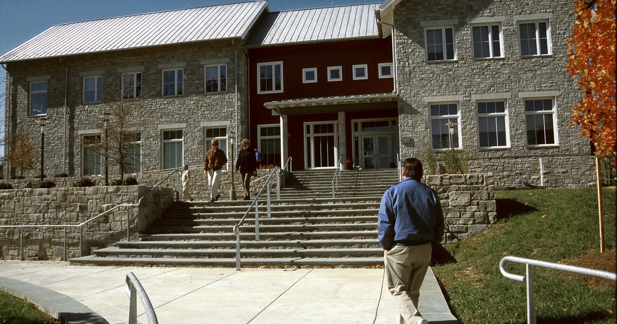 Students walk toward a stone college building with wide front steps on a sunny day