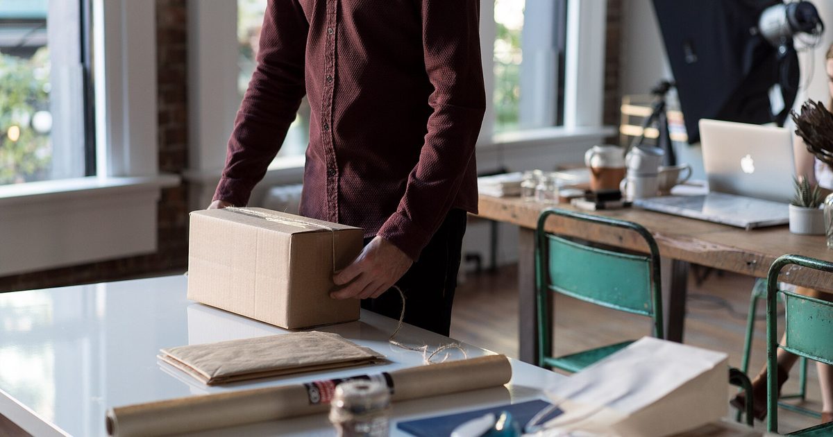 Person in a dark shirt packing a small cardboard box on a white table by a window
