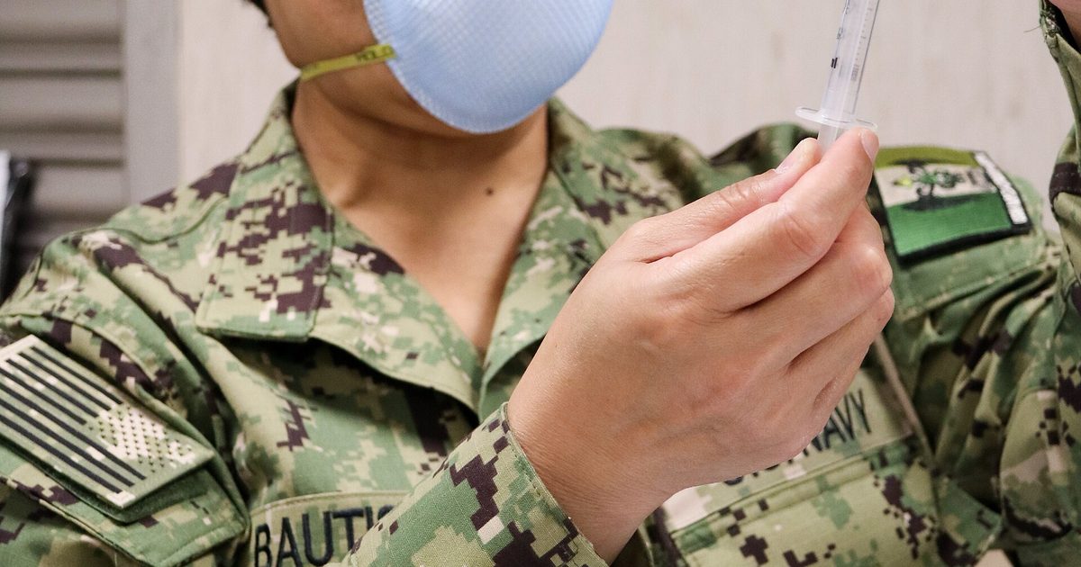 Nurse in scrubs and gloves prepares a vaccine syringe from a vial at a clinic table