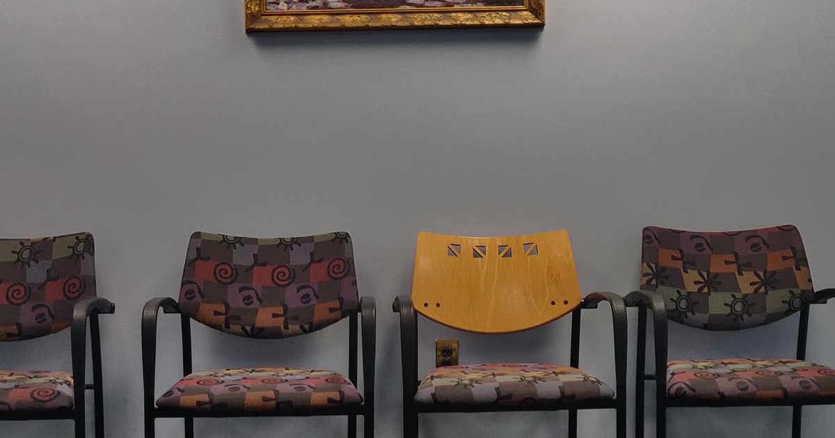A row of empty patterned chairs against a gray wall in a medical waiting room
