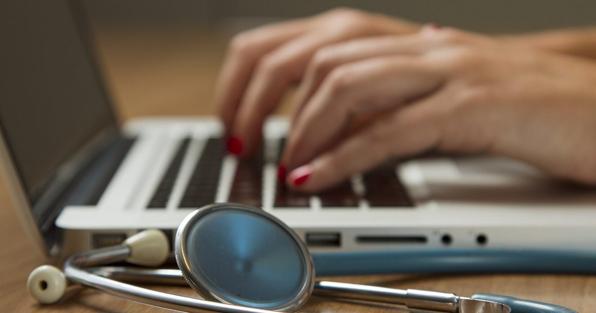 Clinician's hands typing on a laptop with a stethoscope resting on the desk in front