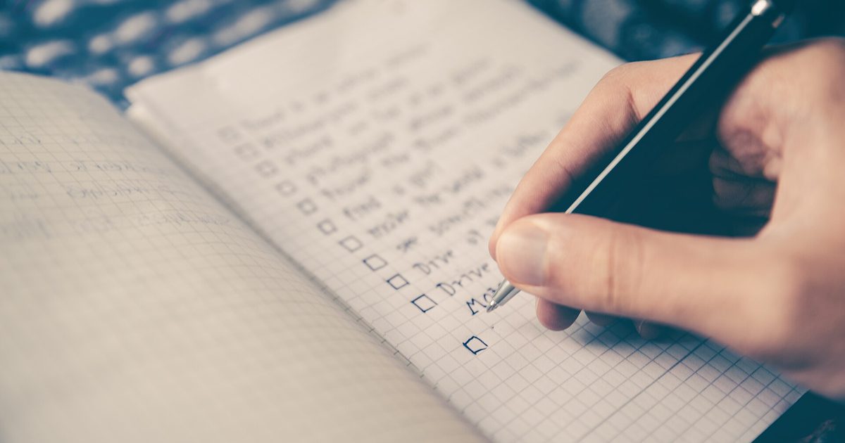 A hand with a pen ticks boxes on a handwritten checklist in a graph-paper notebook